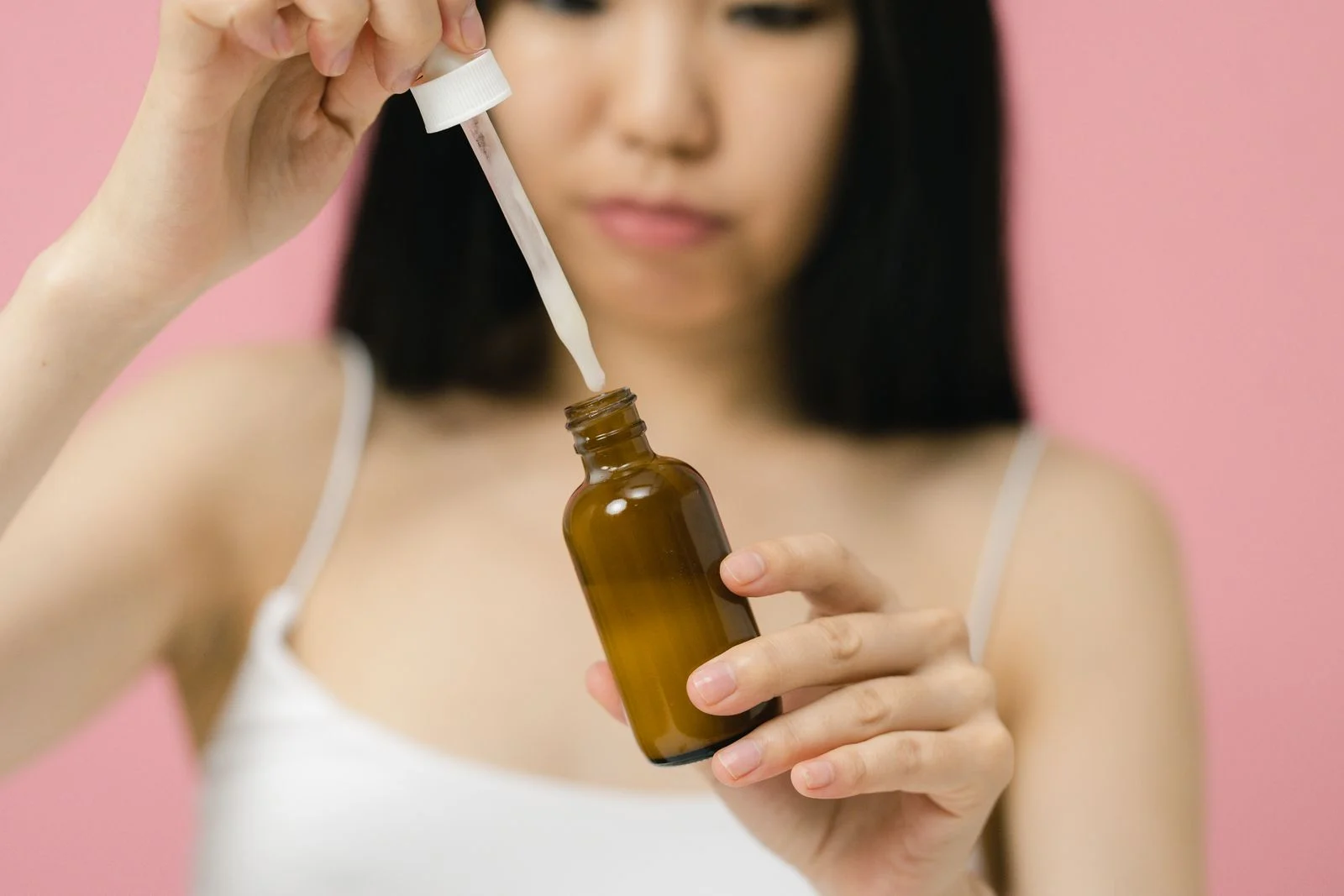 A woman applying skincare serum with a dropper against a pink background
